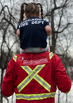 A young girl sitting on the shoulders of a firefighter wearing a Chicago Fire Department shirt and a red Origin and Cause Investigator jacket with reflective stripes.
