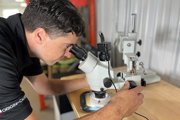 A person in a black shirt is conducting a microscopy experiment, adjusting the focus on a white microscope positioned on a wooden desk with another microscope in the background.